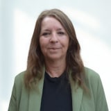 Image of Manager from Vine Lodge Care Home A woman with long brown hair, wearing a green jacket over a black top, stands in front of a plain light background.