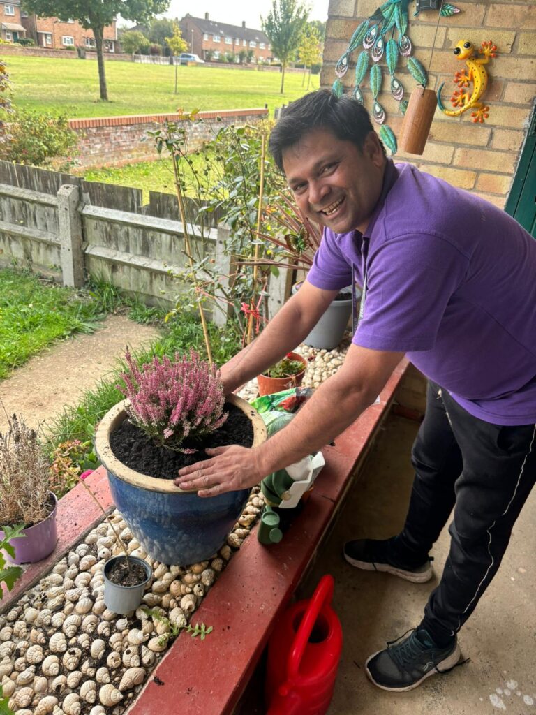 A person in a purple shirt smiles while planting heather in a blue pot on a veranda decorated with seashells, plants, and garden tools.