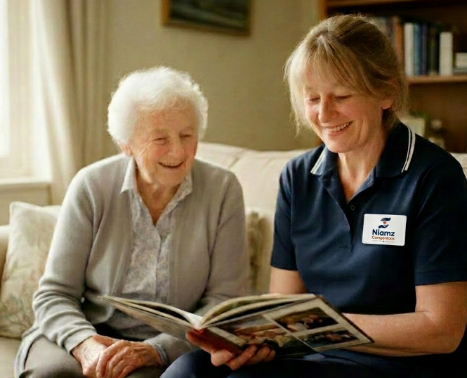An elderly woman and a carer sit together on a sofa, smiling and looking at a photo album. The carer is wearing a navy uniform with a name badge.