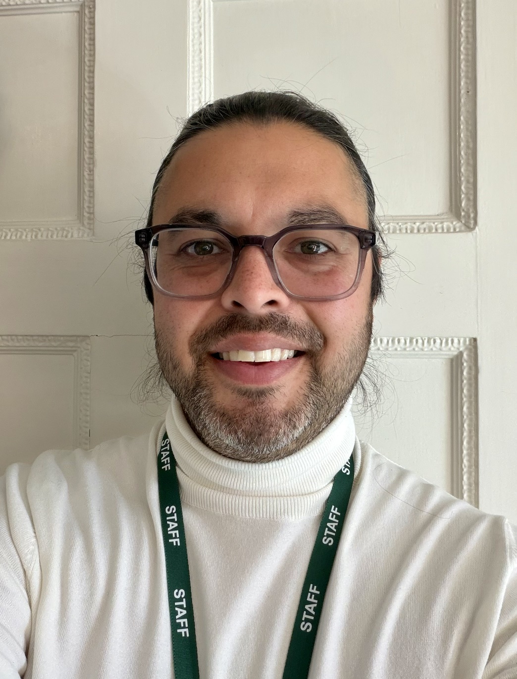 A person wearing glasses, a white polo neck, and a green lanyard labelled STAFF smiles at the camera, standing in front of a white panelled wall.