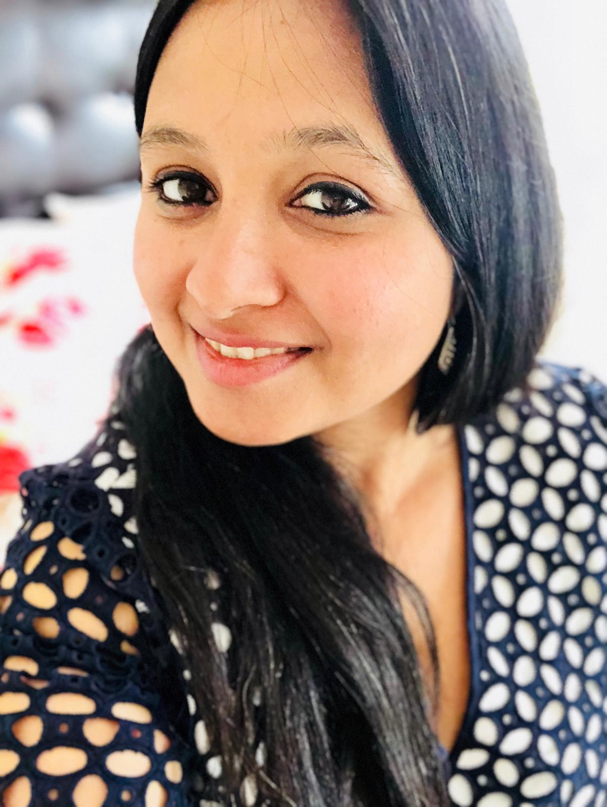 A woman with long black hair and dark eyes smiles at the camera, wearing a navy blue polka dot top and a single earring, with a blurred background.