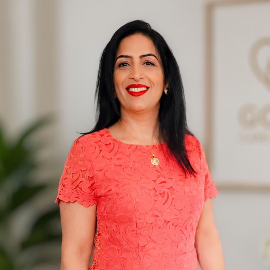 Image of Manager from St David’s Care Home A woman with long dark hair and red lipstick is standing indoors at St David’s Care Home, wearing a coral lace dress and smiling at the camera.