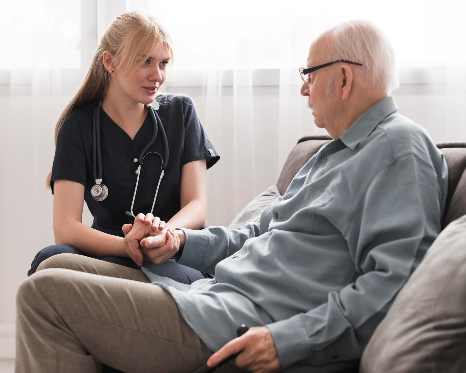 A nurse in navy scrubs sits beside an elderly man on a sofa, holding his hand and listening attentively.