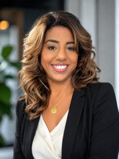 Woman with wavy brown hair, wearing a black blazer and white blouse, smiling in a professional indoor setting with blurred background, reflects the approachable professionalism of Promedica24 Live-in Care Services.